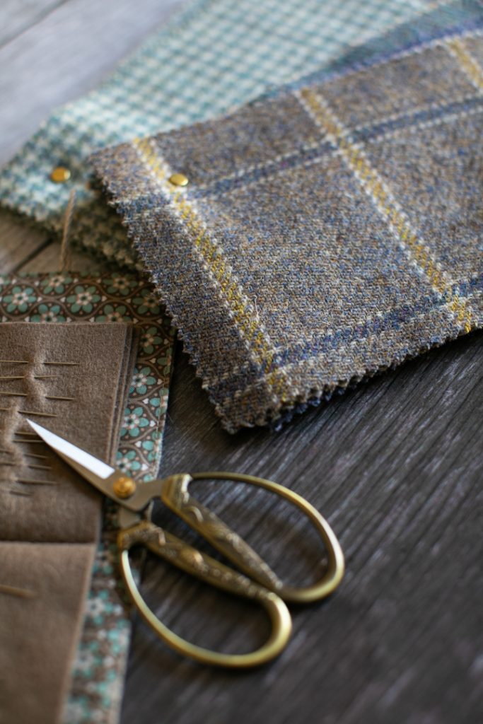 A pile of blue wool fabric samples, a pair of scissors, sewing needles and a fabric tape measure on a wooden table.