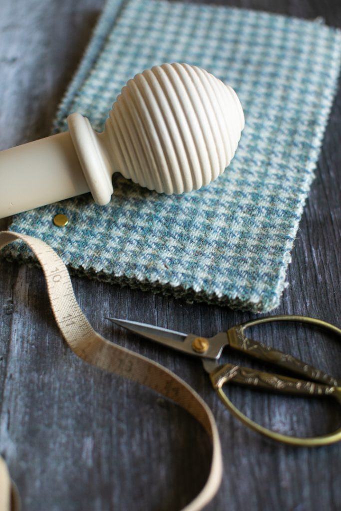 A cream curtain pole next to a blue check pile of wool fabric samples, a pair of scissors and a fabric tape measure on a wooden table.

