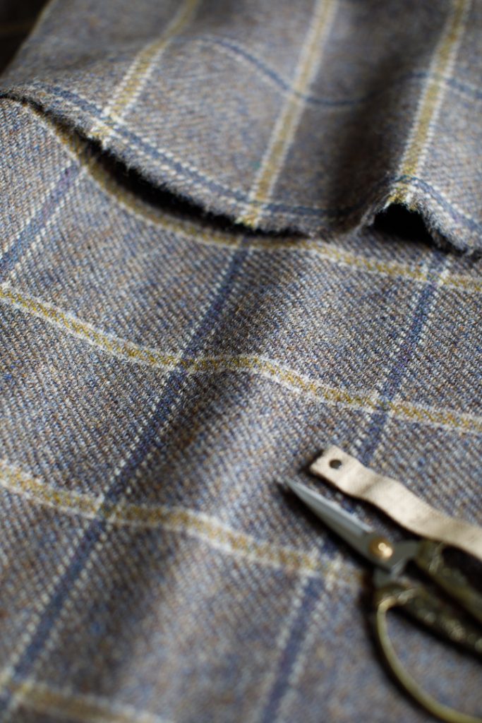 A piece of blue check wool fabric fabric, a pair of scissors and a fabric tape measure on a wooden table.