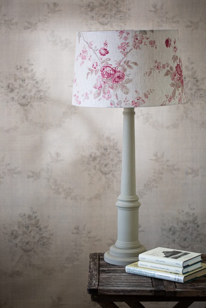 Cream table lamp with a pink floral lampshade, some books on a table and grey and cream wallpaper behind