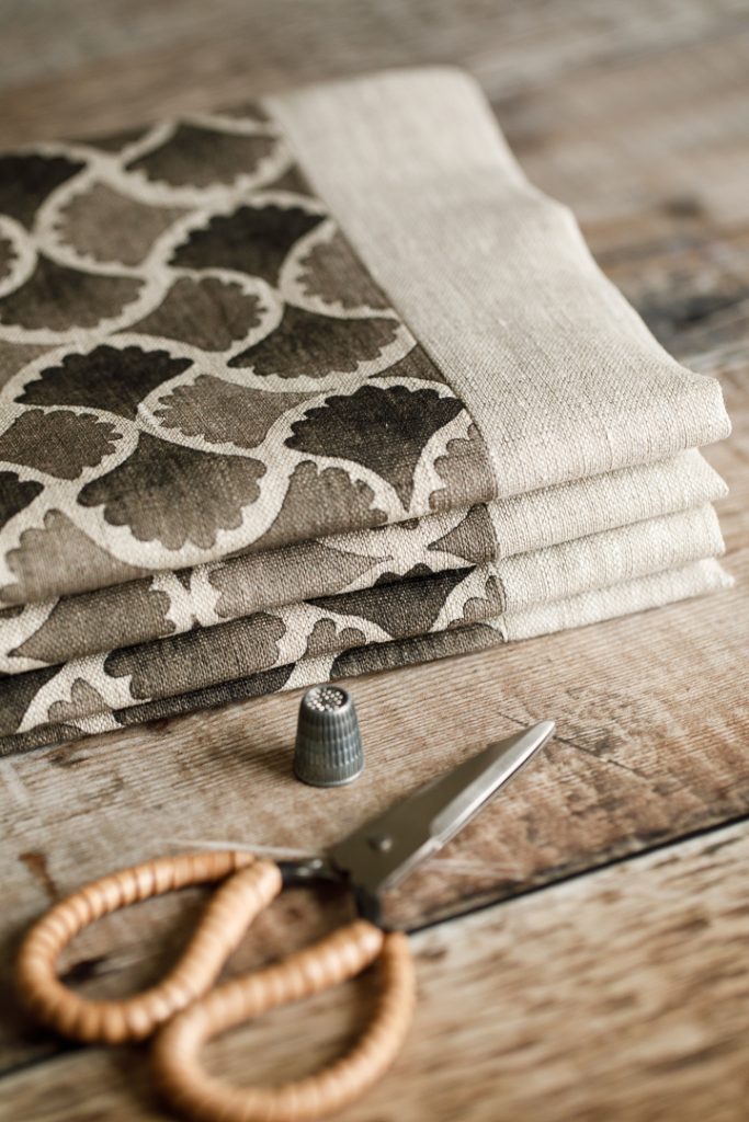 A grey patterned roman blind laying on a wooden table with a pair of scissors and thimble next to it