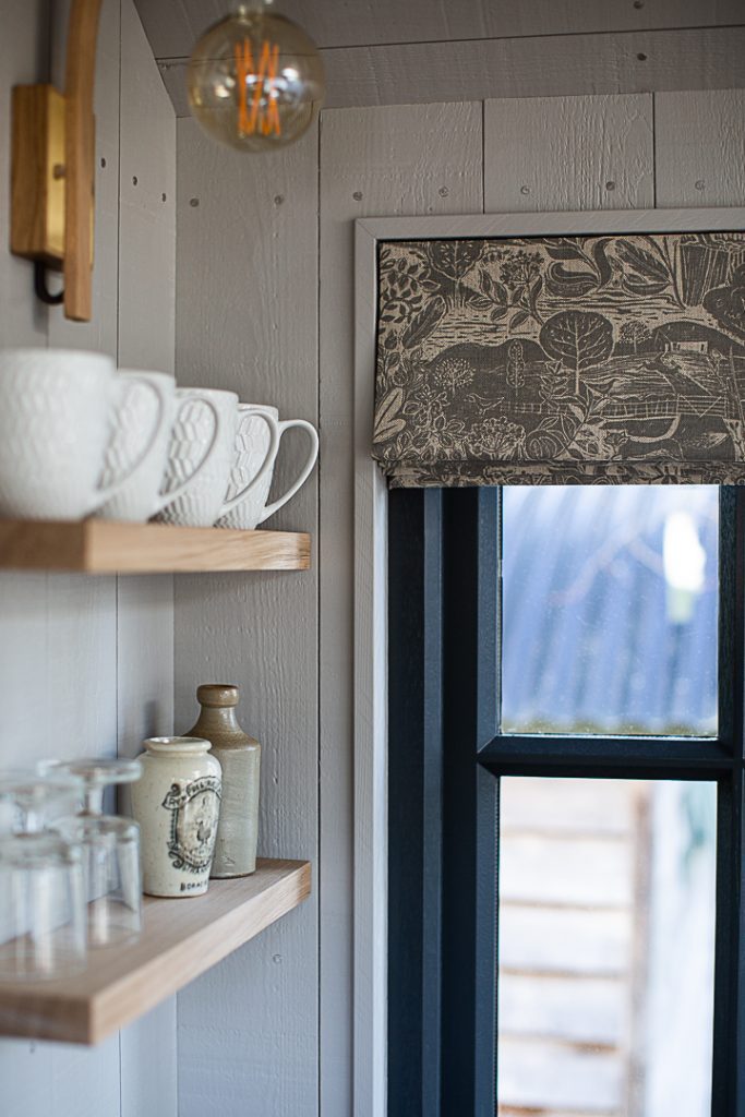 A navy blue window with a grey patterned roman blind, shelves with white mugs and china on